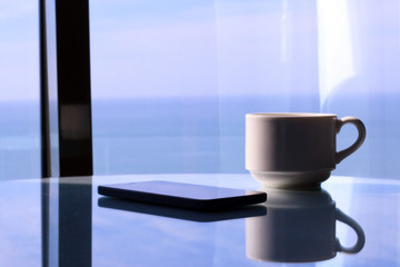  Coffee cup and mobile phone on white table and window with sea view. 