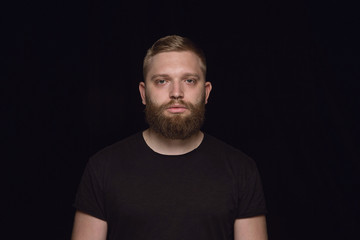 Fototapeta premium Close up portrait of young caucasian man isolated on black studio background. Photoshot of real emotions of male model. Standing and looks serious. Facial expression, human nature and emotions concept