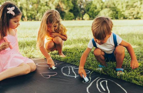 Children Sitting On The Green Grass Playing With Colorful Chalks Happy Little Kids Drawing With Chalks In The Park Friends Boy And Two Girls Having Fun On Sunlight Outdoor Childhood And Friendship