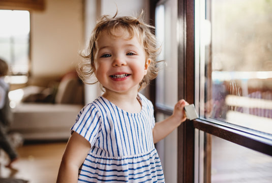 A toddler girl standing by window indoors at home, looking at camera.