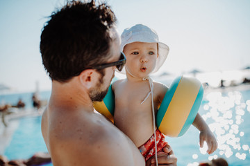 Father with small child with armbands standing by swimming pool on summer holiday.