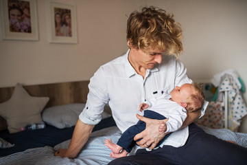 A young father with a newborn baby son sitting on bed at home.