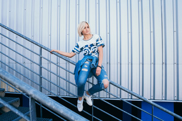 Beautiful girl with short white hair dressed in jeans in urban style sits on the background of a white wall. Place for text