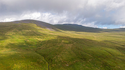 A panoramic aerial view of a mountain with grassy green slope, stream and stormy summit under a majestic srormy white clouds