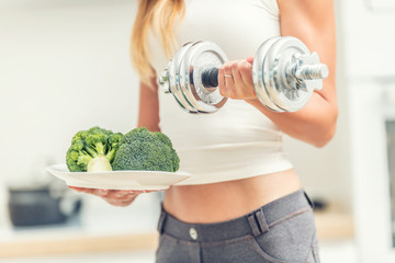Young woman with sports figure in the kitchen holding dumbbells in her hands and a plate of broccoli