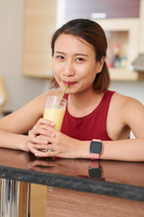 Obraz na płótnie Canvas Portrait of young smiling Asian woman drinking sweet tasty fruit cocktail and looking at camera