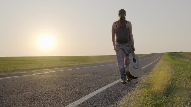 Girl traveler with guitar walking along country road at sunset alone.