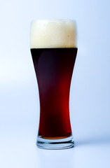A full glass of dark beer with a large cap of froth. Slightly bluish background. Side view. Close-up.