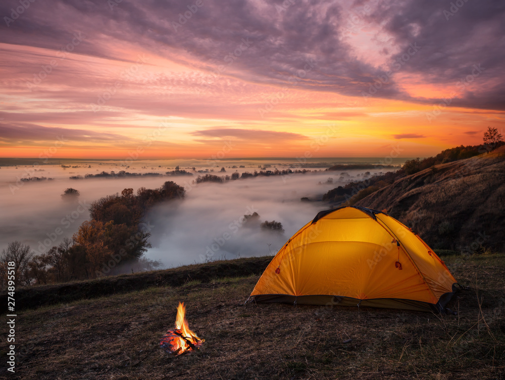 Wall mural orange lit inside tent and fire over misty river at sunset