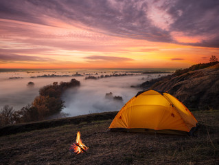 Orange lit inside tent and fire over misty river at sunset
