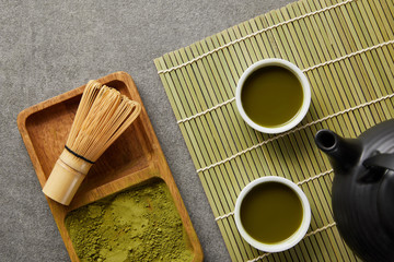 top view of green matcha powder with bamboo whisk in wooden board near teapot and white cups with green tea on table mat
