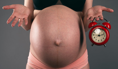 Pregnant woman is holding in hands a red alarm clock on a gray background. Waiting for the baby.
