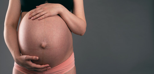 Young pregnant woman is holding her belly by her hands on a gray background with copy space.