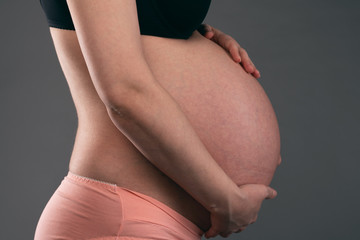 Young pregnant woman is holding her belly by her hands on a gray background.