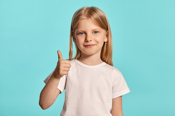 Close-up studio shot of a lovely blonde little girl in a white t-shirt posing against a blue...