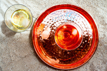 Glasses of aperol spritz and white wine with ice and fruits, copper tray on sand background outdoors