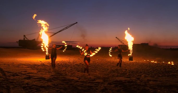 Fire show three women in their hands twist burning spears and fans in the sand with a man with two flamethrowers in slow motion.