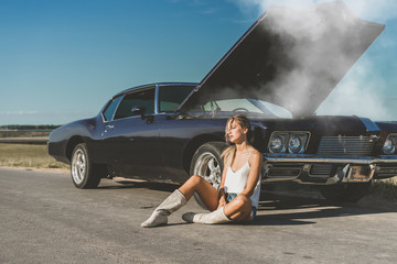 Young female sits near overheated car in the field, bright sunlight, steam under the hood