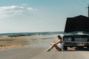 Young female sits near overheated car in the field, bright sunlight, steam under the hood