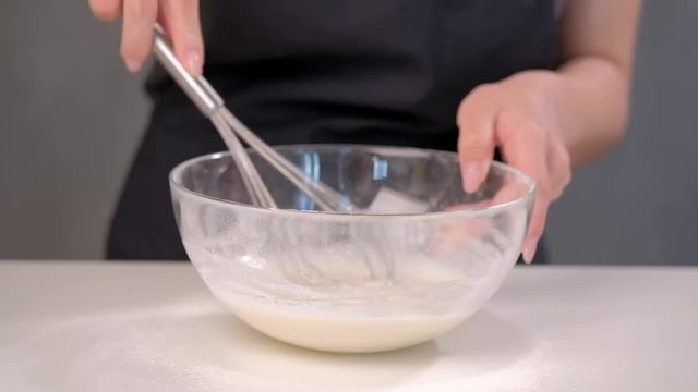 A female cook pastry chef whips dough in a glass bowl with a metal whisk. Close-up.