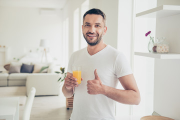 Close up photo attractive he him his homey guy arm hand orange juice glass energy refreshment thumb up symbol advising good quality white nightwear t-shirt bright kitchen flat room indoors