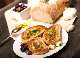 Sliced traditional toast bread with olive oil, olives, garlic and herbs on a plate and dark table