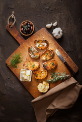 Sliced traditional toast bread with butter, olives, garlic and herbs on a cutting board, napkin and dark table