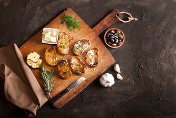Sliced traditional toast bread with butter, olives, garlic and herbs on a cutting board, napkin and dark table