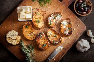 Fototapeta premium Sliced traditional toast bread with butter, olives, garlic and herbs on a cutting board and dark table