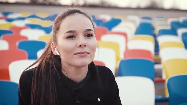 A young and beautiful brunette girl, sitting in the spectator row of a sports arena, watches the training of athletes and listens to music through wireless headphones.