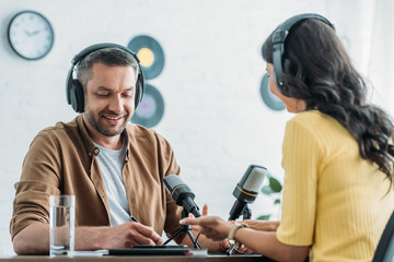 cheerful radio host in headphones writing in notebook while sitting at workplace near colleague