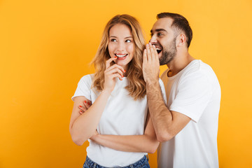 Image of happy couple man and woman in basic t-shirts whispering secrets or gossips to each other - obrazy, fototapety, plakaty