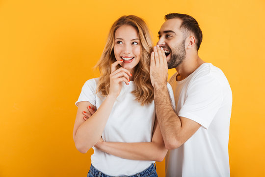 Image of happy couple man and woman in basic t-shirts whispering secrets or gossips to each other