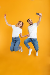 Full length image of cheerful couple rejoicing and jumping while taking selfie photo on cellphone