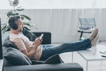 adult man in earphones sitting on sofa with legs on table and using smartphone