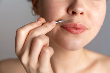 A young woman plucks her hair over her upper lip with tweezers. The concept of getting rid of...