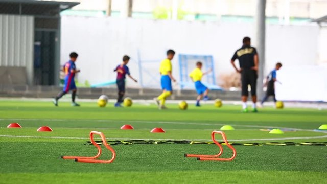 orange hurdles on green artificial turf with blurry coach and kid soccer are training. kid soccer players are jogging and control ball. soccer equipment in football academy.