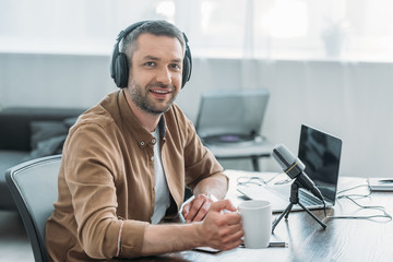 handsome radio host smiling at camera while sitting at workplace with cup of coffee