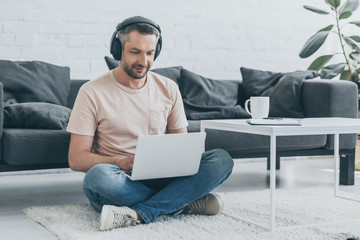 handsome man in headphones sitting on floor with crossed legs and using laptop