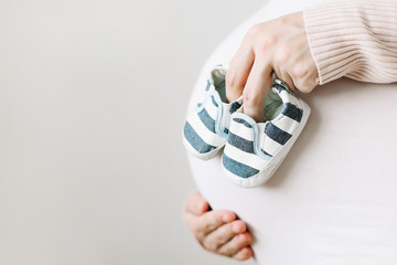 Belly of a pregnant woman in white clothes, mother hands holding shoes for the newborn baby