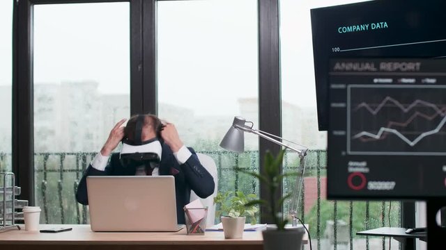 Businessman in formal suit puts ON a VR headset. Modern office with big windows. Rainy day