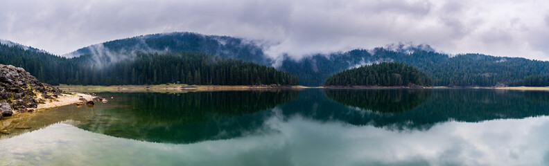 Obraz na płótnie Canvas Montenegro, XXL landscape panorama of glassy calm water of black lake reflecting green forest and hills in foggy atmosphere in popular durmitor national park near zabljak