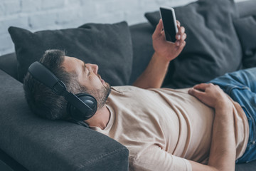adult man in headphones holding smartphone with blank screen while resting on sofa