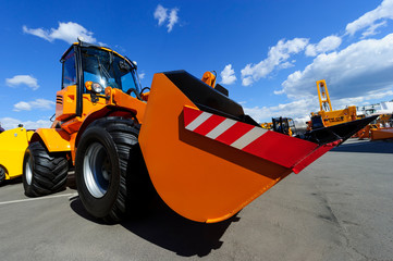 Wheel loader, orange construction machine with big scoop, heavy industry, blue sky and white clouds on background