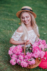 Fototapeta premium Beautiful girl in vintage dress and straw hat sitting on grass and holding pink peonies in rustic basket