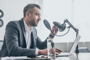 handsome radio host speaking in microphone while sitting at workplace near laptop and glass of water