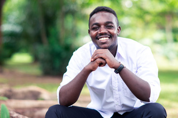 African business man in white shirt smiling and sitting outside with green trees on background.