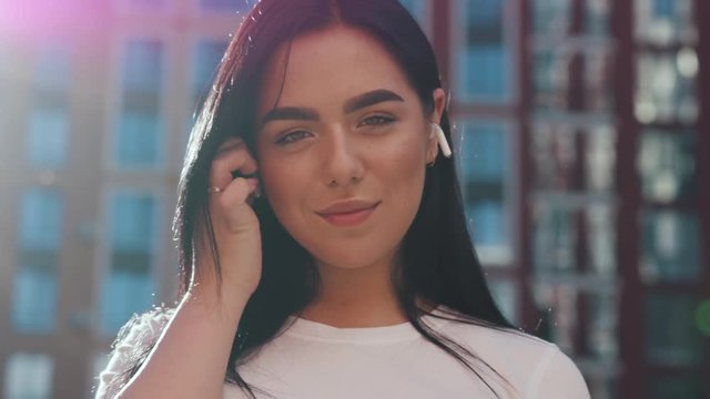 Close up view portrait of happy woman putting on headphones, earpods outdoors in bright sunlight. Sun beams, enjoying music, having fun. Modern gadgets, technology concept