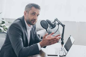 handsome radio host gesturing and looking at camera while sitting at workplace in broadcasting...