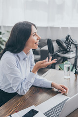 smiling radio host speaking in microphone while sitting at desk near laptop in broadcasting studio